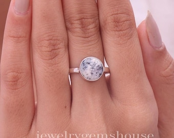 Puede incluir: Un anillo de plata con una piedra redonda engastada en bisel, con una base blanca e inclusiones negras moteadas. La piedra tiene una superficie lisa y pulida, centrada en una banda de plata fina. El texto jewelrygemshouse es visible en la parte inferior.