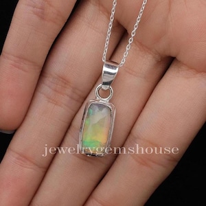 Peut inclure: Un pendentif en opale rectangulaire serti dans une monture en argent poli, suspendu à une fine chaîne en argent. L'opale présente des reflets irisés verts, jaunes et orange. Le texte jewelrygemshouse est visible sur la partie inférieure.
