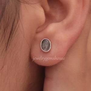 May include: A pair of silver stud earrings with a dark gray oval-shaped gemstone. The earrings are shown in a person's ear.