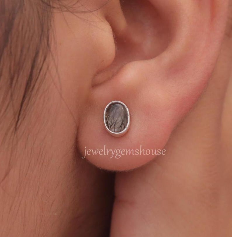 May include: A pair of silver stud earrings with a dark gray oval-shaped gemstone. The earrings are shown in a person's ear.