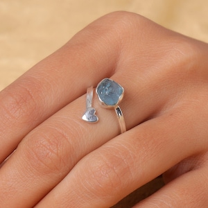 Könnte beinhalten: Ein silberner Ring mit einem rohen, hellblauen Edelstein. Der Ring hat ein offenes Design mit einem kleinen Herzdetail. Der Edelstein ist unregelmäßig geformt und in einer einfachen Lünette gefasst.