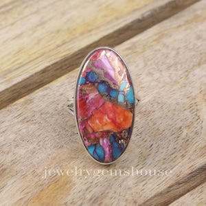 May include: An oval-shaped ring featuring a large, colourful gemstone set in sterling silver. The gemstone is a mix of pink, orange, blue, and brown, with a swirling pattern.