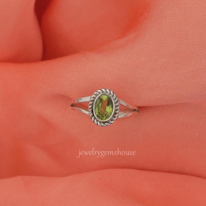May include: A silver ring featuring an oval-cut light green peridot gemstone set within a twisted rope-style metal border. The band is smooth and tapers toward the back. The text jewelrygemshouse is visible in the lower center of the image.