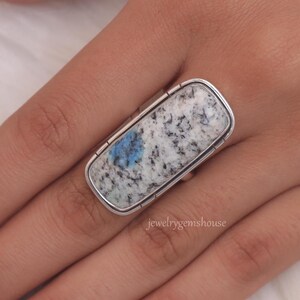 May include: A silver ring with a rectangular blue and white stone. The stone has a speckled pattern and a blue spot in the center. The ring is on a person's finger.