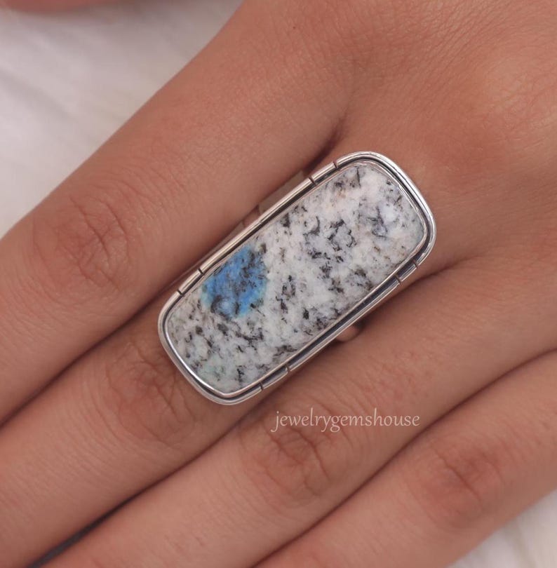 May include: A silver ring with a rectangular blue and white stone. The stone has a speckled pattern and a blue spot in the center. The ring is on a person's finger.