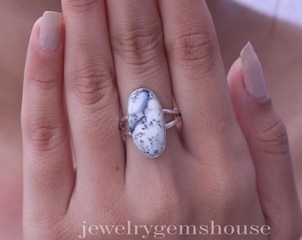 Puede incluir: Un anillo con una piedra preciosa de ópalo dendrítico de forma ovalada engastada en una banda de plata. La piedra es blanca con inclusiones oscuras en forma de helecho en toda su superficie. La banda de metal tiene un diseño dividido.