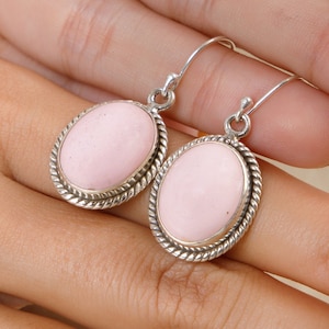 May include: A pair of oval-shaped earrings featuring smooth, light pink stones set in polished silver frames. Each earring is surrounded by a decorative twisted rope border and hangs from a simple silver hook wire for secure attachment.