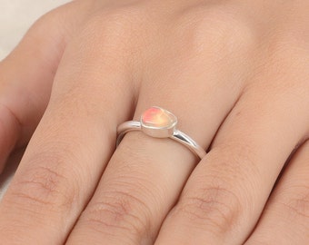 Pode incluir: Um anel de prata com uma única pedra preciosa de opala lisa, engastada em bisel, com reflexos iridescentes de laranja e rosa. A banda é polida, fina e arredondada, exibindo um design simples que destaca o jogo de cores natural da pedra.