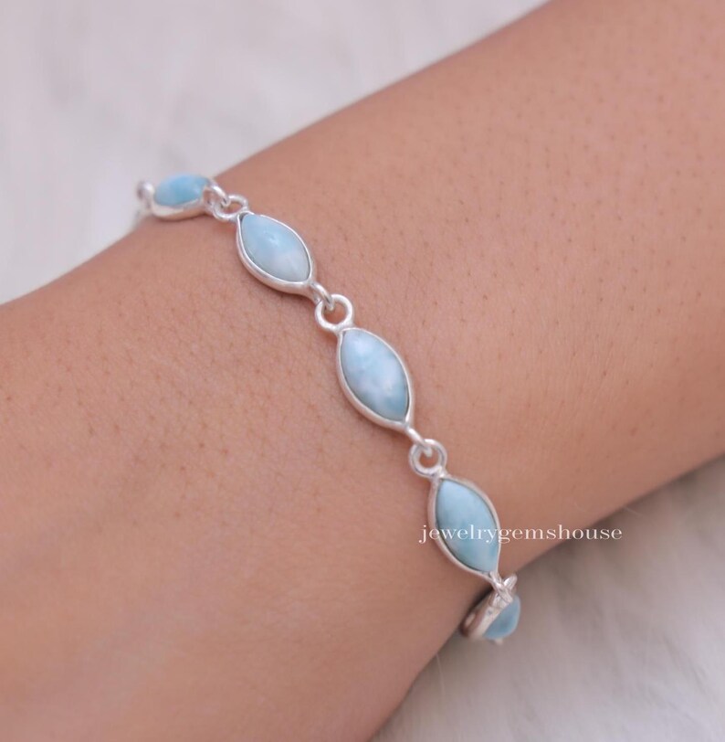 Puede incluir: Una pulsera de plata con piedras preciosas ovaladas de color azul claro. Las piedras est&aacute;n unidas por peque&ntilde;os conectores de plata. La pulsera se lleva en la mu&ntilde;eca. El texto "jewelrygemshouse" es visible.