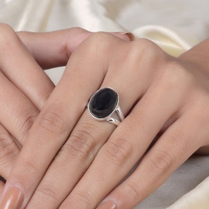Puede incluir: Un anillo de plata con una gran piedra preciosa negra de forma ovalada. El anillo tiene un diseño de caña dividida, con la banda dividiéndose en dos partes al acercarse a la piedra preciosa. El anillo se muestra en una mano.