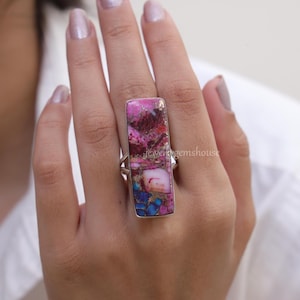 Op de afbeelding: Een zilveren ring met een rechthoekige roze, blauwe en gouden edelsteen. De edelsteen heeft een marmerpatroon.