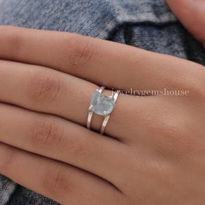 Peut inclure: Bague en argent ornée d'une pierre précieuse bleu clair de forme irrégulière. La bague présente un design à tige fendue, avec deux bandes parallèles qui se rejoignent au niveau de la pierre précieuse. Le texte "jewelrygemshouse" est visible.
