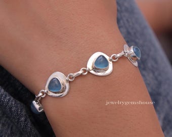 May include: A silver bracelet featuring blue gemstone accents. The bracelet has a series of linked, open-backed settings, each holding a triangular or teardrop-shaped blue stone. The bracelet is worn on a wrist, with the jewelrygems house logo visible.