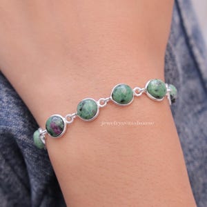 Pode incluir: Uma pulseira de prata com pedras preciosas verdes e rosas. A pulseira tem um total de sete pedras preciosas, cada uma com um padrão único. A pulseira está no pulso de uma pessoa.
