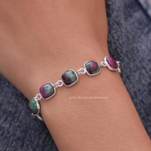 May include: A silver bracelet featuring square-shaped green and pink gemstones. The bracelet is worn on a person's wrist.