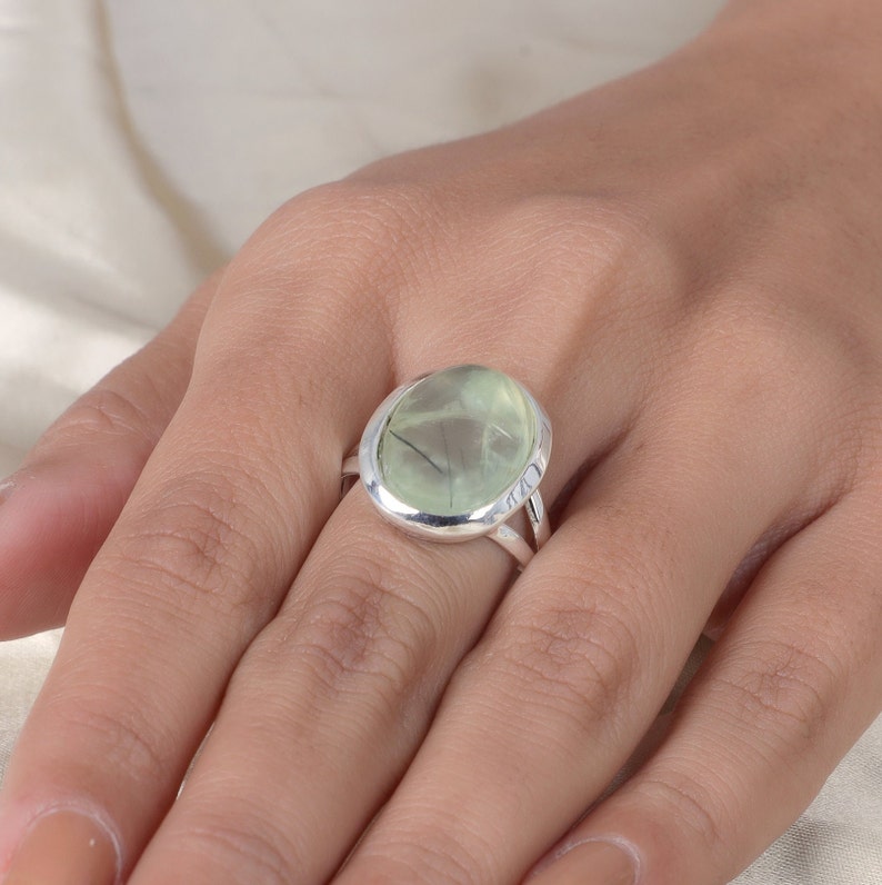 Peut inclure: Une bague en argent orn&eacute;e d'une grande pierre pr&eacute;cieuse ovale de couleur vert p&acirc;le, pr&eacute;sentant des inclusions sombres en forme d'aiguilles. La pierre est sertie dans une monture en argent lisse et poli, avec un anneau &agrave; double branche.
