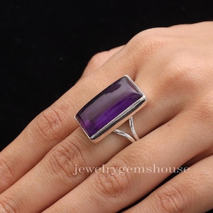 May include: A rectangular amethyst ring with a silver band. The large, polished purple gemstone is set in a silver bezel. The ring is on a hand, with the text "jewelrygemshouse" visible.