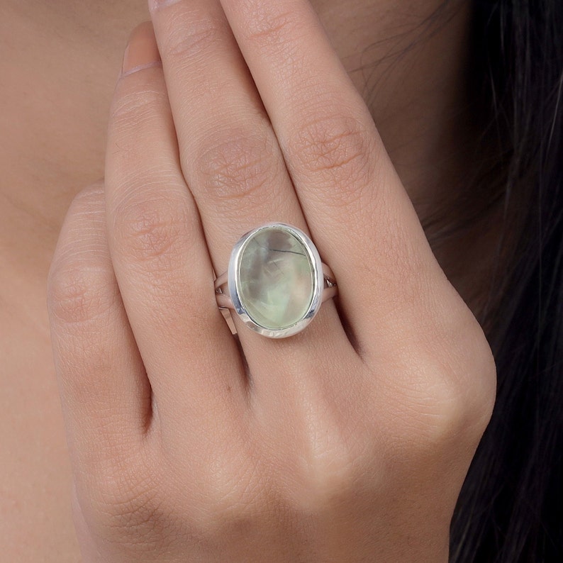 Peut inclure: Une bague en argent orn&eacute;e d'une grande pierre pr&eacute;cieuse ovale, translucide et vert p&acirc;le, pr&eacute;sentant de fines inclusions sombres. La pierre est sertie dans un chaton en argent poli et lisse, avec un anneau &agrave; double branche qui s'affine.
