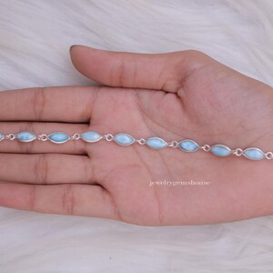 Puede incluir: Un brazalete de plata con piedras preciosas ovaladas de color azul claro. El brazalete se muestra en una mano, destacando la delicada cadena y las piedras pulidas. La joya se encuentra sobre un fondo blanco suave.