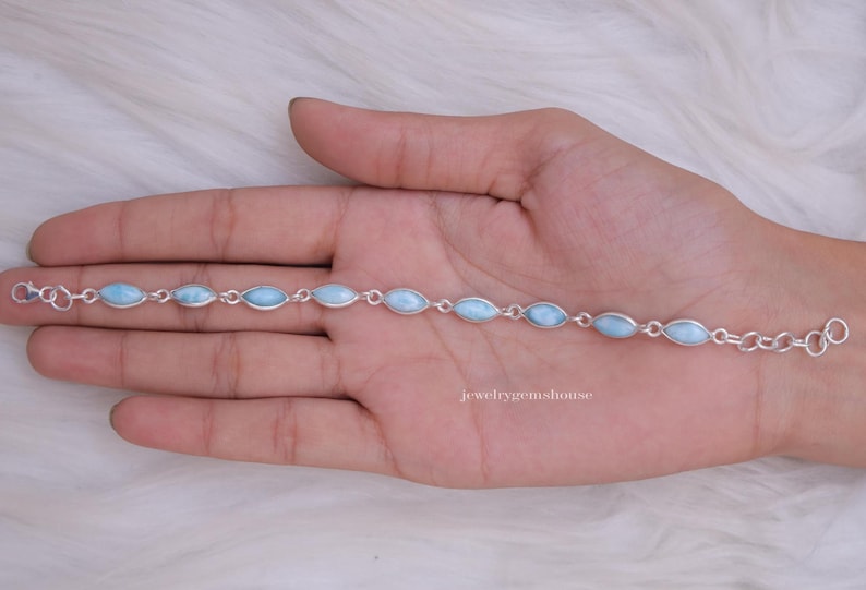 Puede incluir: Un brazalete de plata con piedras preciosas ovaladas de color azul claro. El brazalete se muestra en una mano, destacando la delicada cadena y las piedras pulidas. La joya se encuentra sobre un fondo blanco suave.