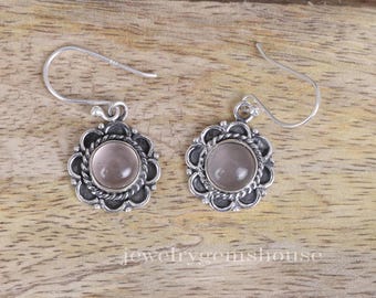 May include: A pair of silver-toned earrings with a floral design. Each earring features a round, light pink gemstone set within a decorative, scalloped frame. The earrings have a hook closure.