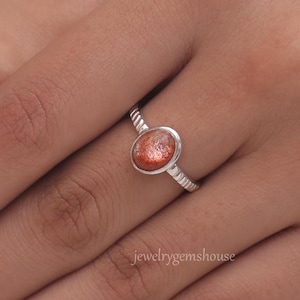 Puede incluir: Un anillo de plata con una piedra preciosa de piedra de sol. El anillo tiene una banda texturizada y la piedra preciosa de forma ovalada es de color naranja cálido con motas rojas y doradas.