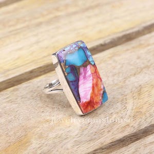 May include: A silver ring featuring a rectangular, multi-coloured gemstone. The gemstone displays shades of pink, orange, blue, and purple.