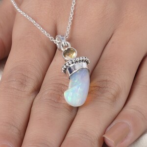 Puede incluir: Un colgante de plata con una piedra de &oacute;palo blanco iridiscente curvada, engastada en una tapa de plata con cuentas y rematada con una peque&ntilde;a piedra preciosa de citrino amarillo redonda y facetada. El colgante cuelga de una fina cadena.