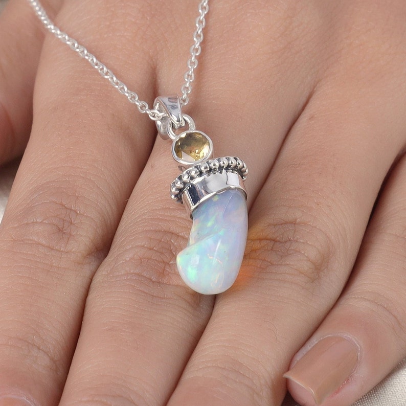 Puede incluir: Un colgante de plata con una piedra de &oacute;palo blanco iridiscente curvada, engastada en una tapa de plata con cuentas y rematada con una peque&ntilde;a piedra preciosa de citrino amarillo redonda y facetada. El colgante cuelga de una fina cadena.