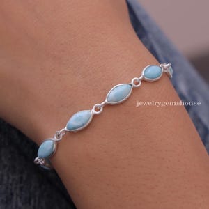 Puede incluir: Una pulsera de plata con piedras preciosas ovaladas de color azul claro. La pulsera est&aacute; conectada por peque&ntilde;os eslabones de plata y se lleva en la mu&ntilde;eca. El texto "jewelrygemshouse" es visible.