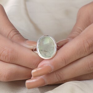 Peut inclure: Une bague en argent orn&eacute;e d'une grande pierre cabochon ovale vert p&acirc;le. La pierre pr&eacute;sente des inclusions sombres en forme d'aiguilles visibles sur toute sa surface translucide. L'anneau en m&eacute;tal est lisse et poli, encadrant la pierre.