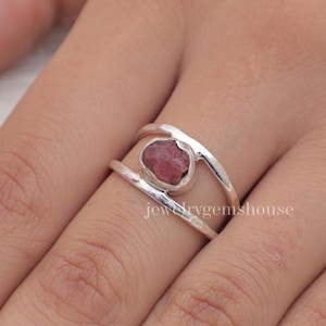 May include: A silver ring with a pink gemstone set in a double band design. The ring is on a finger.