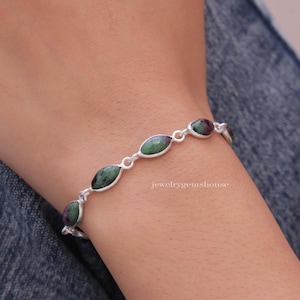 May include: A silver bracelet featuring green and pink oval-shaped gemstones. The bracelet is worn on a person's wrist.