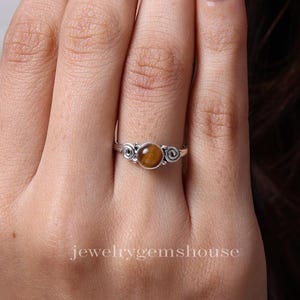 May include: A silver ring featuring a brown tiger's eye gemstone. The ring has a simple design with two swirls on either side of the stone.