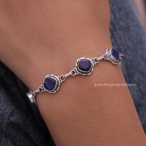 Può includere: Un bracciale a catena in argento con quattro pietre preziose sfaccettate blu scuro di forma quadrata, incastonate in castoni d'argento ossidato con bordi decorativi. Il testo jewelrygemshouse è visibile sul lato destro del bracciale.