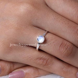 Könnte beinhalten: Ein Silberring mit einem einzelnen, runden, durchscheinenden Mondstein in einer glatten Fassung. Das Band ist dünn und poliert und zeigt ein schlichtes Design, das die irisierende Qualität des Edelsteins in der Mitte des Fingers hervorhebt.