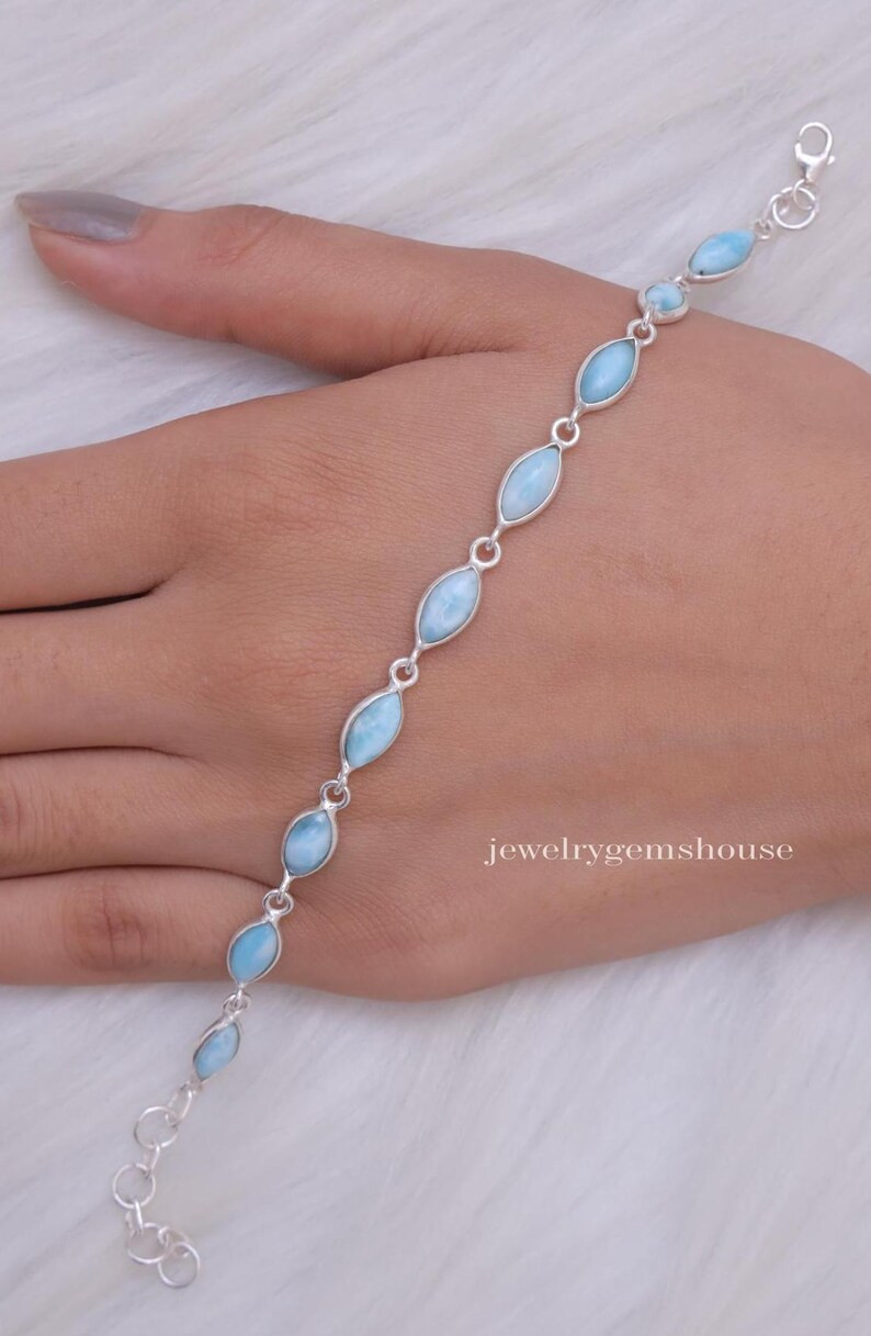 Puede incluir: Una pulsera de plata con piedras preciosas ovaladas de color azul claro. La pulsera est&aacute; conectada por peque&ntilde;os eslabones de plata y tiene un cierre de broche. La joya se muestra en una mano, con el texto "jewelrygemshouse" visible.