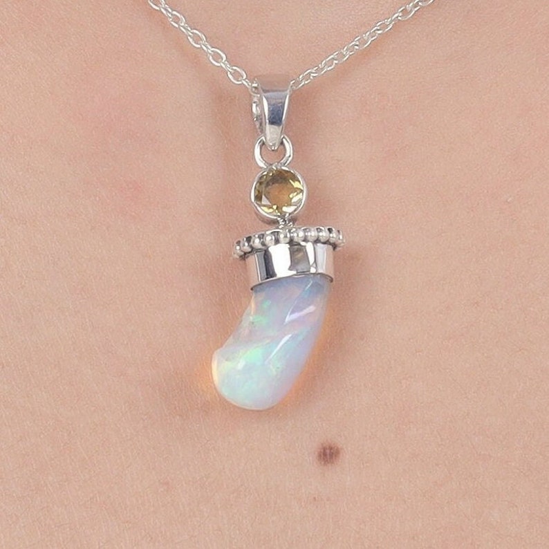 Puede incluir: Un collar con colgante de plata que presenta una piedra de &oacute;palo blanco iridiscente curvada, engastada en una tapa de plata con borde de cuentas. Sobre el &oacute;palo, una peque&ntilde;a piedra preciosa redonda de color amarillo parduzco.