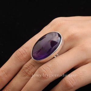 May include: A silver ring featuring a large, oval amethyst gemstone. The deep purple stone is set in a silver band. The ring is on a person's finger. The text "jewelrygemshouse" is visible at the bottom of the image.