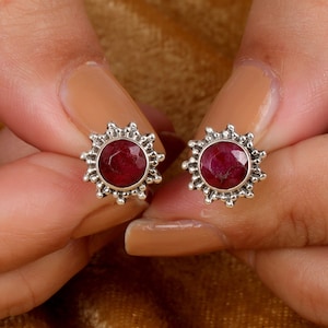 Può includere: Un paio di orecchini a lobo in argento con una pietra preziosa di rubino rosso al centro di ogni orecchino. Gli orecchini hanno un design a raggiera attorno alla pietra preziosa.