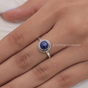 Pode incluir: Um anel de prata com uma pedra preciosa redonda de lápis-lazúli azul profundo, engastada numa borda de metal trançado. A banda é lisa e polida, e o texto jewelrygemshouse é visível no lado direito da cravação do anel.