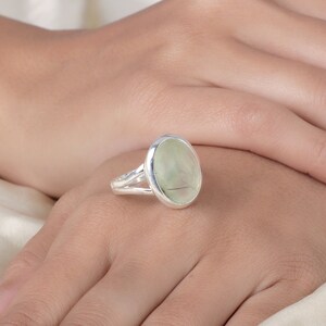 Peut inclure: Une bague en argent orn&eacute;e d'une grande pierre pr&eacute;cieuse ovale de couleur vert p&acirc;le avec des inclusions sombres visibles. La pierre est sertie dans un chaton en argent poli et lisse, et l'anneau est form&eacute; de deux fins fils d'argent parall&egrave;les.