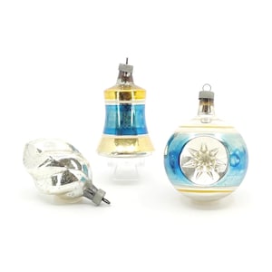 May include: Three vintage Christmas ornaments. One is a silver teardrop shape, another is a blue, gold, and white bell shape, and the third is a blue and white globe with a star design. All have metal hanging loops.