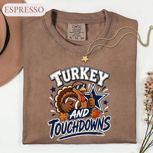 May include: An espresso-colored t-shirt featuring a graphic of a turkey wearing sunglasses, holding a football, and giving a thumbs up. The text reads "TURKEY AND TOUCHDOWNS" in blue and white lettering, accompanied by a blue star and orange maple leaves.