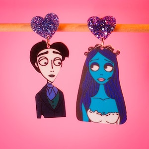 May include: A pair of earrings featuring a cartoon image of a man and a woman. The man is wearing a suit and the woman is wearing a white dress. The earrings are decorated with glitter hearts.