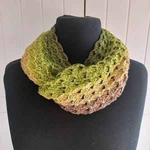 Könnte beinhalten: Ein gehäkelter Infinity-Schal mit einem Farbverlauf, der von hellem Olivgrün zu mittlerem Salbeigrün übergeht und mit einem tiefen, erdigen Braunton endet. Das Accessoire weist durchgehend ein strukturiertes, offenes Häkelmuster auf.
