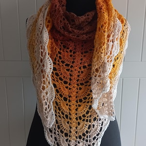 Könnte beinhalten: Ein dreieckiges Häkeltuch mit einem komplizierten fächerartigen Stichmuster. Das Accessoire zeigt einen Farbverlauf, der von einem tiefen Dunkelbraun am Halsbereich über Orange- und Gelbtöne bis hin zu einem hellen Cremeweiß an den Rändern reicht.