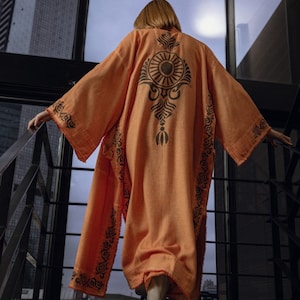 Pode incluir: Uma mulher vestindo um quimono longo laranja com um design geom&eacute;trico preto e branco. O quimono tem uma silhueta larga e fluida e &eacute; decorado com padr&otilde;es intrincados. A mulher est&aacute; em uma escada, olhando para fora de uma janela.