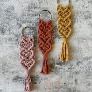 May include: Three macrame keyrings in pink, rust, and mustard yellow. Each keyring has a woven design, a metal ring at the top, and a tassel at the bottom. They are displayed against a textured, neutral background.