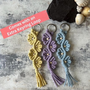 May include: Three macrame keychains in yellow, lavender, and light blue, each featuring a flower-like pattern with a central wooden bead and a tassel at the bottom. Each keychain includes a metal ring and a pink label stating Comes with an Extra Keyring Loop.